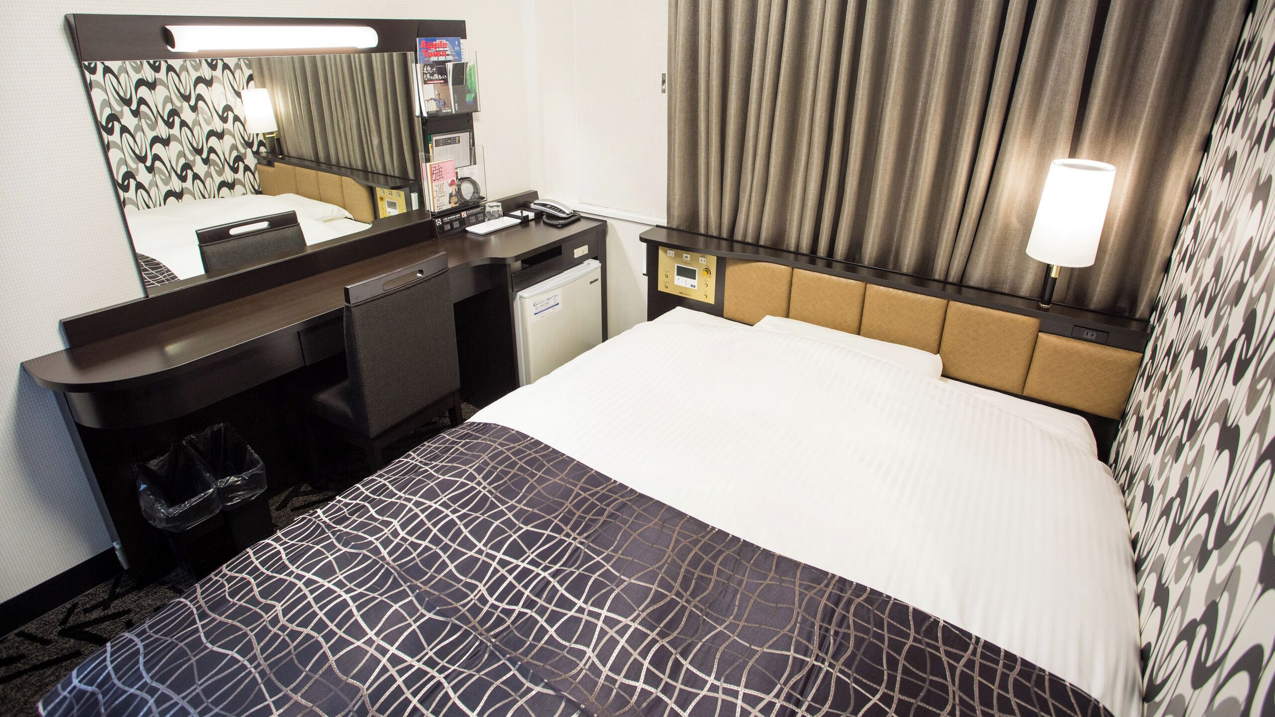 A hotel room with a bed, a desk, a mirror, a telephone, and a small refrigerator.