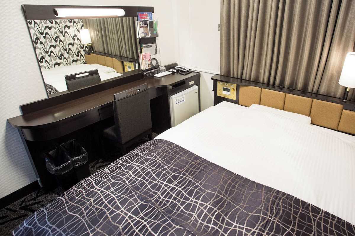 A hotel room with a bed, a desk, a mirror, a telephone, and a small refrigerator.