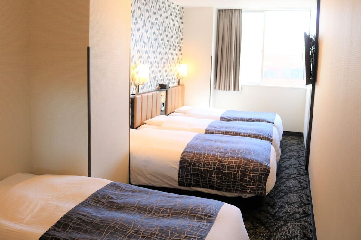 A hotel room with two beds, a patterned wall, a window with curtains, and a television mounted on the wall.