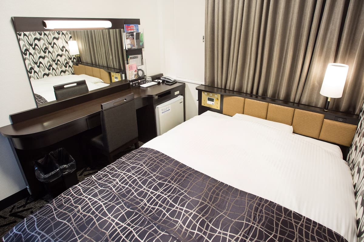 A hotel room with a bed, a desk, a chair, a mirror, a nightstand, a lamp, and a window with curtains.