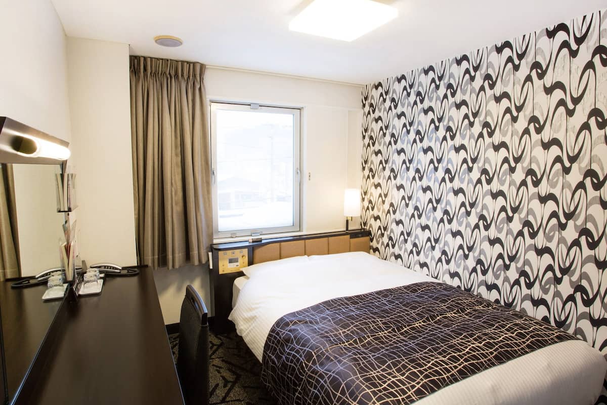 A hotel room with a bed, a desk, a chair, a window, and a patterned wall.