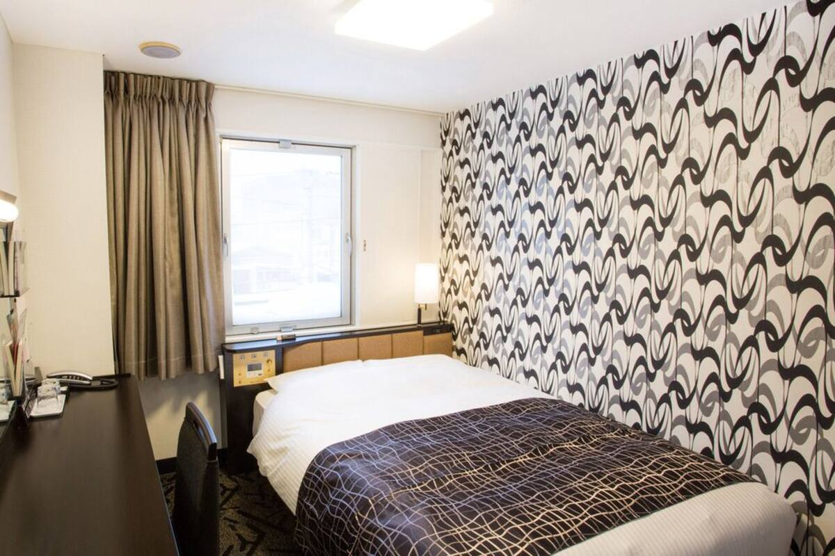 Une chambre d’hôtel avec un lit, un bureau doté d’un miroir, une fenêtre avec des rideaux et un mur orné d’un motif.
