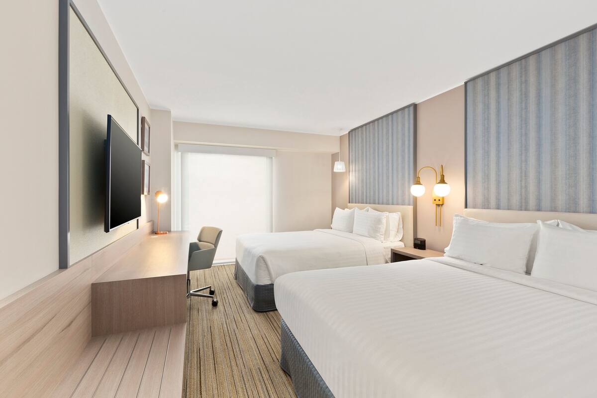A hotel room with two beds, a desk, a chair, a TV, and a large window.