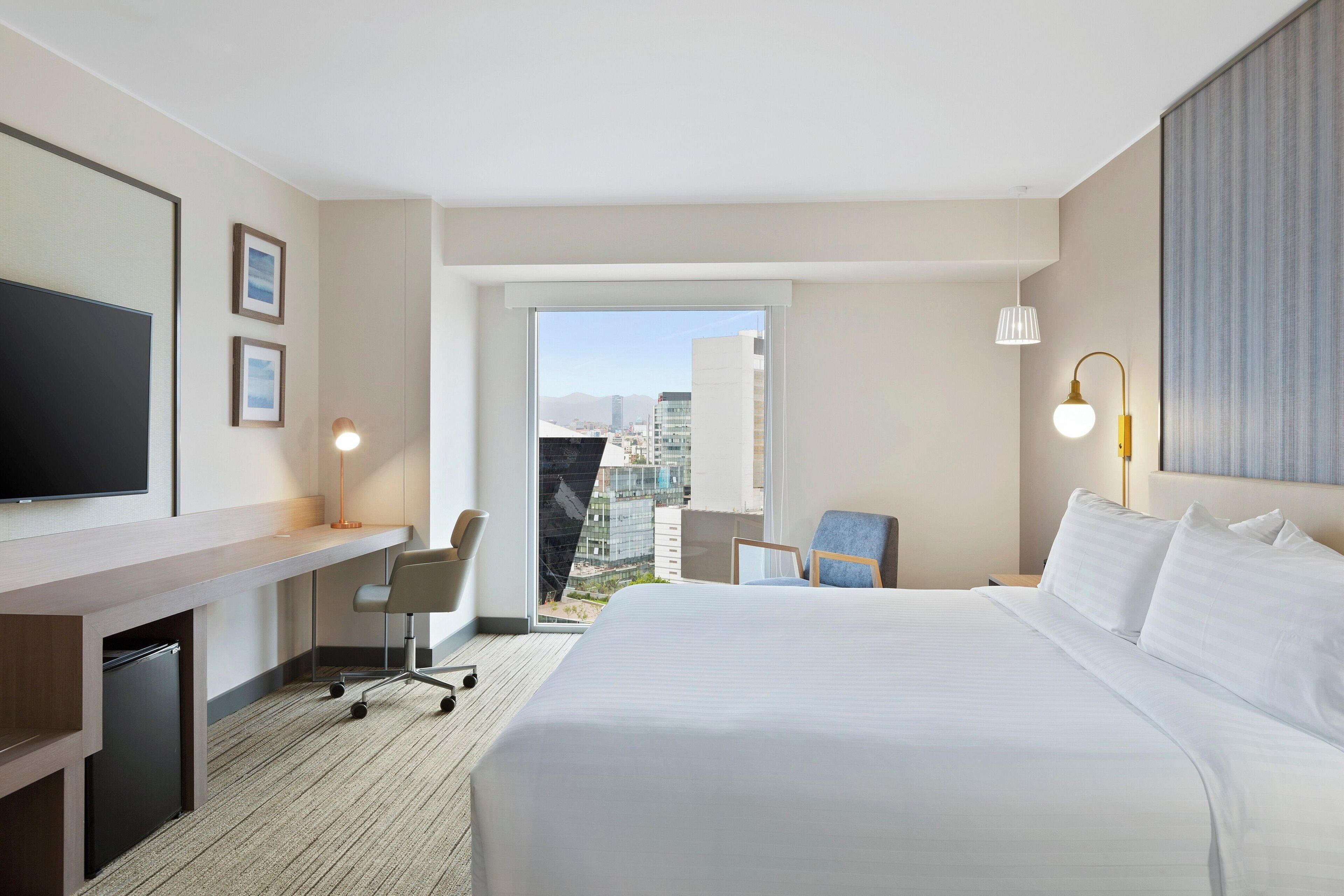 A hotel room with a bed, desk, chair, TV, and a view of the city.