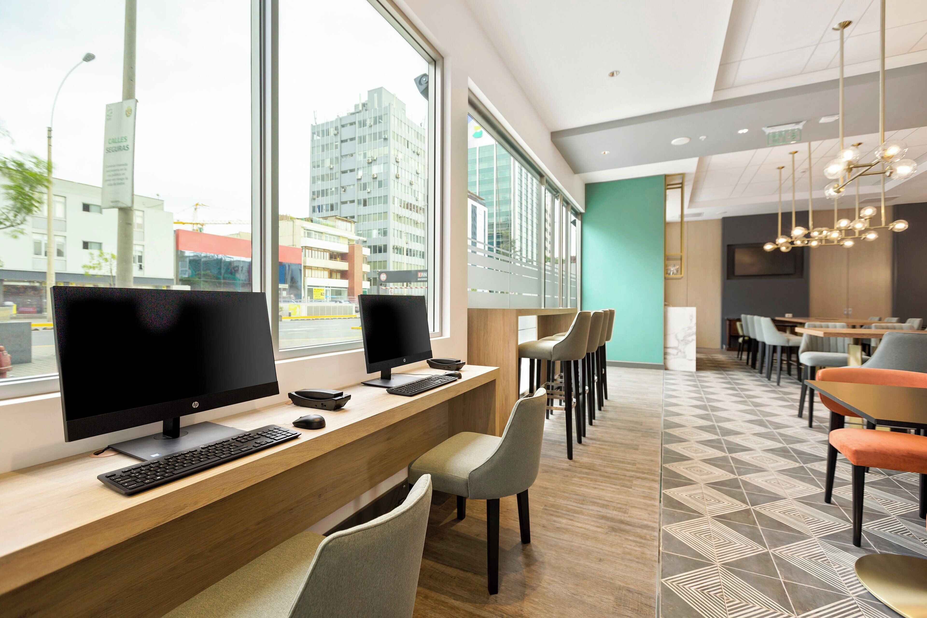 A modern office space with two HP monitors, a keyboard, and a mouse on a wooden desk. There are multiple chairs and a dining area with tables and chairs.