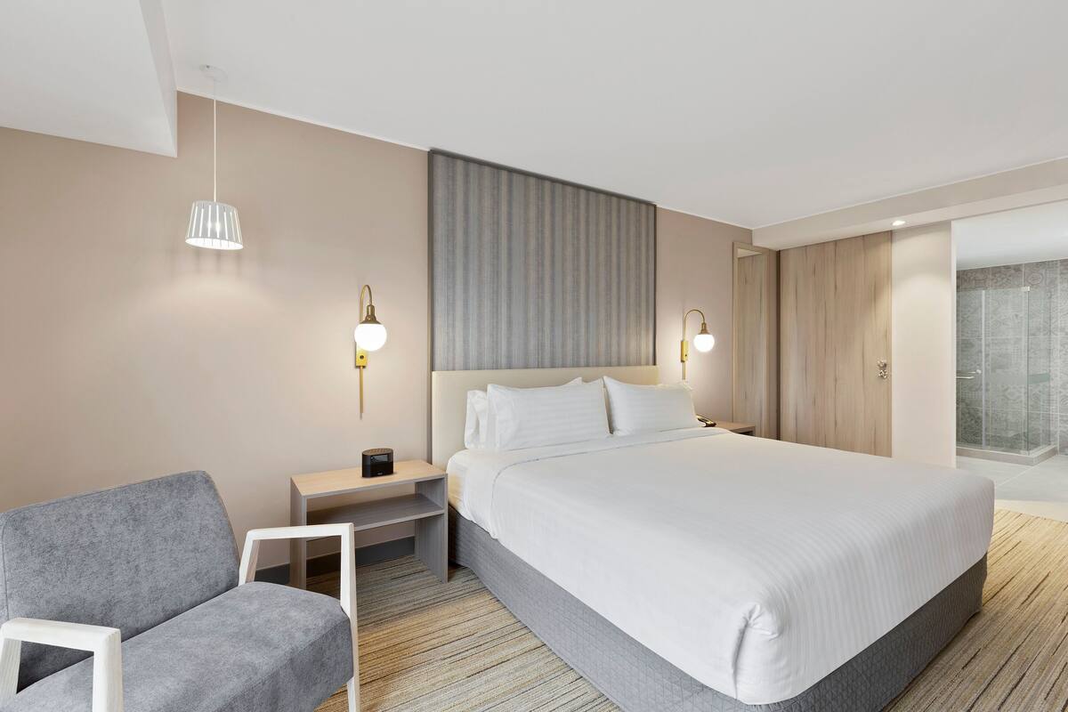 A modern hotel room with a large bed, a grey armchair, bedside tables with lamps, and a large window with a view of a cityscape.