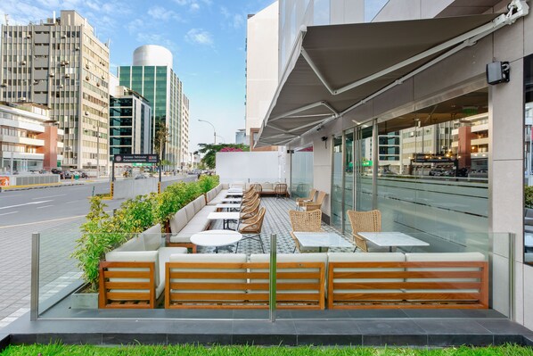 Un espace extérieur aménagé avec des bancs et des tables en bois, entouré de verdure et donnant sur une rue bordée d’immeubles.