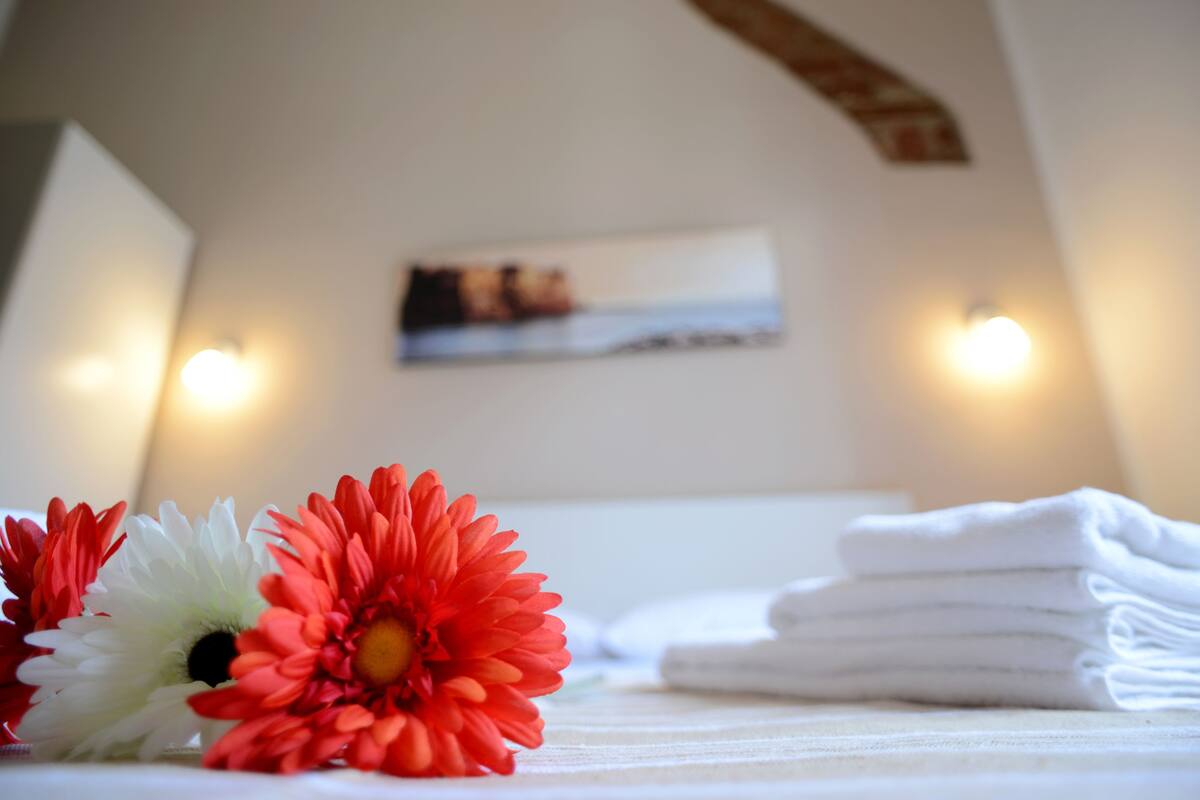 A neatly made bed with folded white towels, a red and white flower, and a painting on the wall.