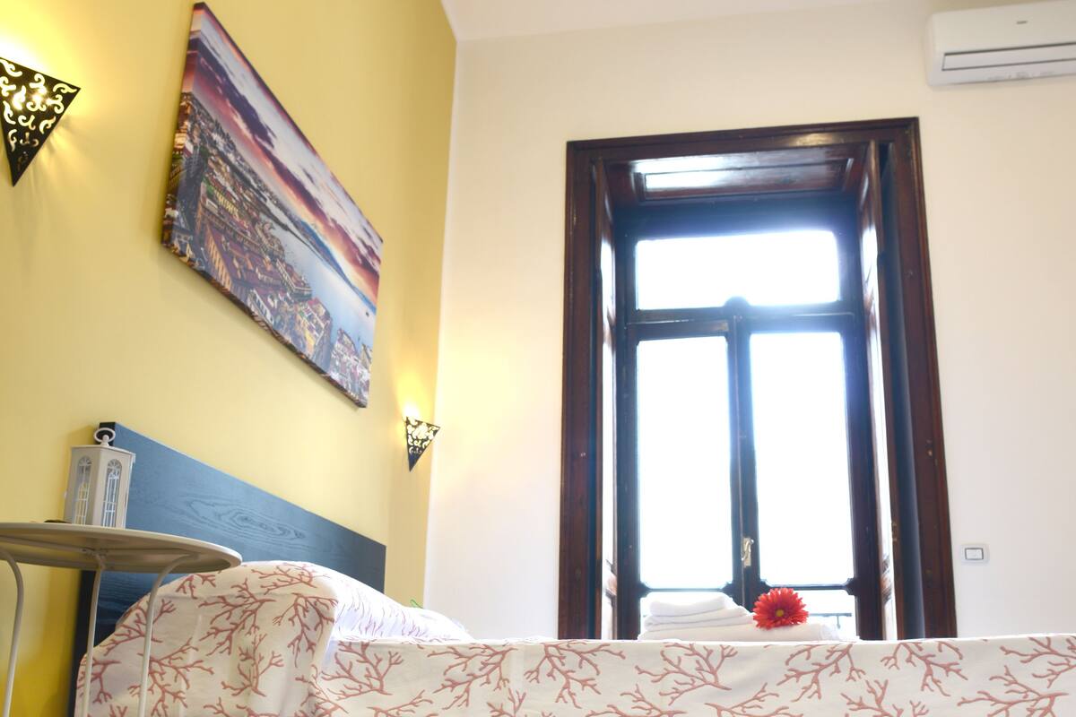 A room with a bed, a window, a painting on the wall, and a small table with a lamp.