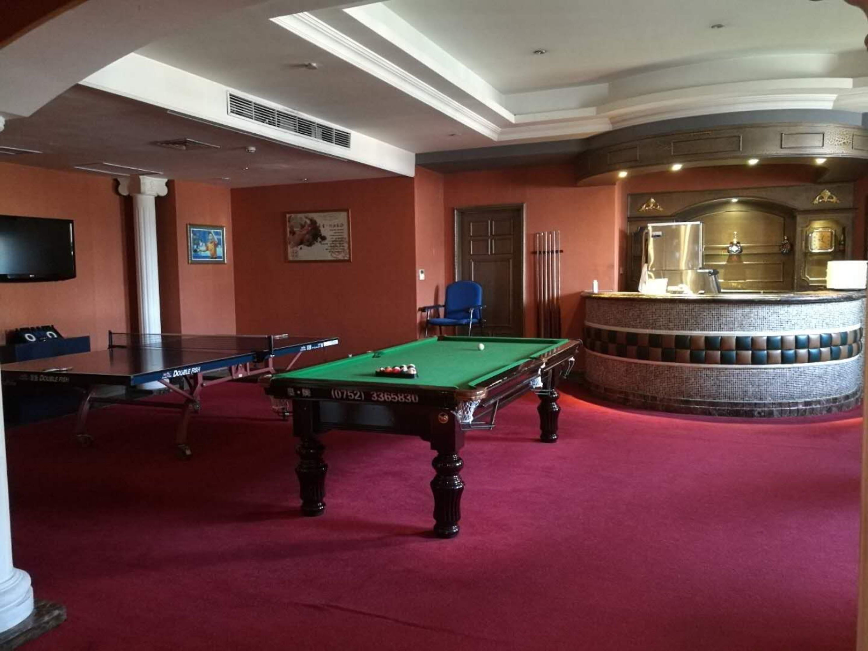 Una mesa de billar en una sala con alfombra roja, una zona de bar con estampado de cuadros y un televisor colgado en la pared.