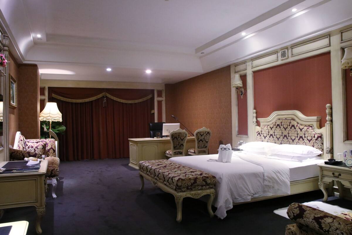A hotel room with a large bed, a desk, a chair, a bench, and a desk lamp.