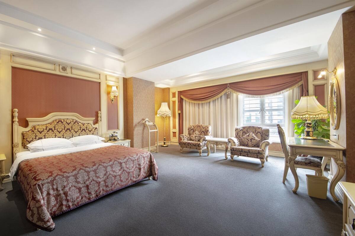 A spacious hotel room with a large bed, two armchairs, a small table, and a desk with a lamp.