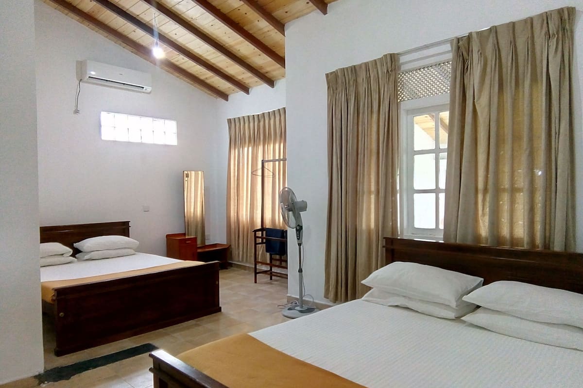 A hotel room with two beds, a standing fan, a window with curtains, and a ceiling fan.