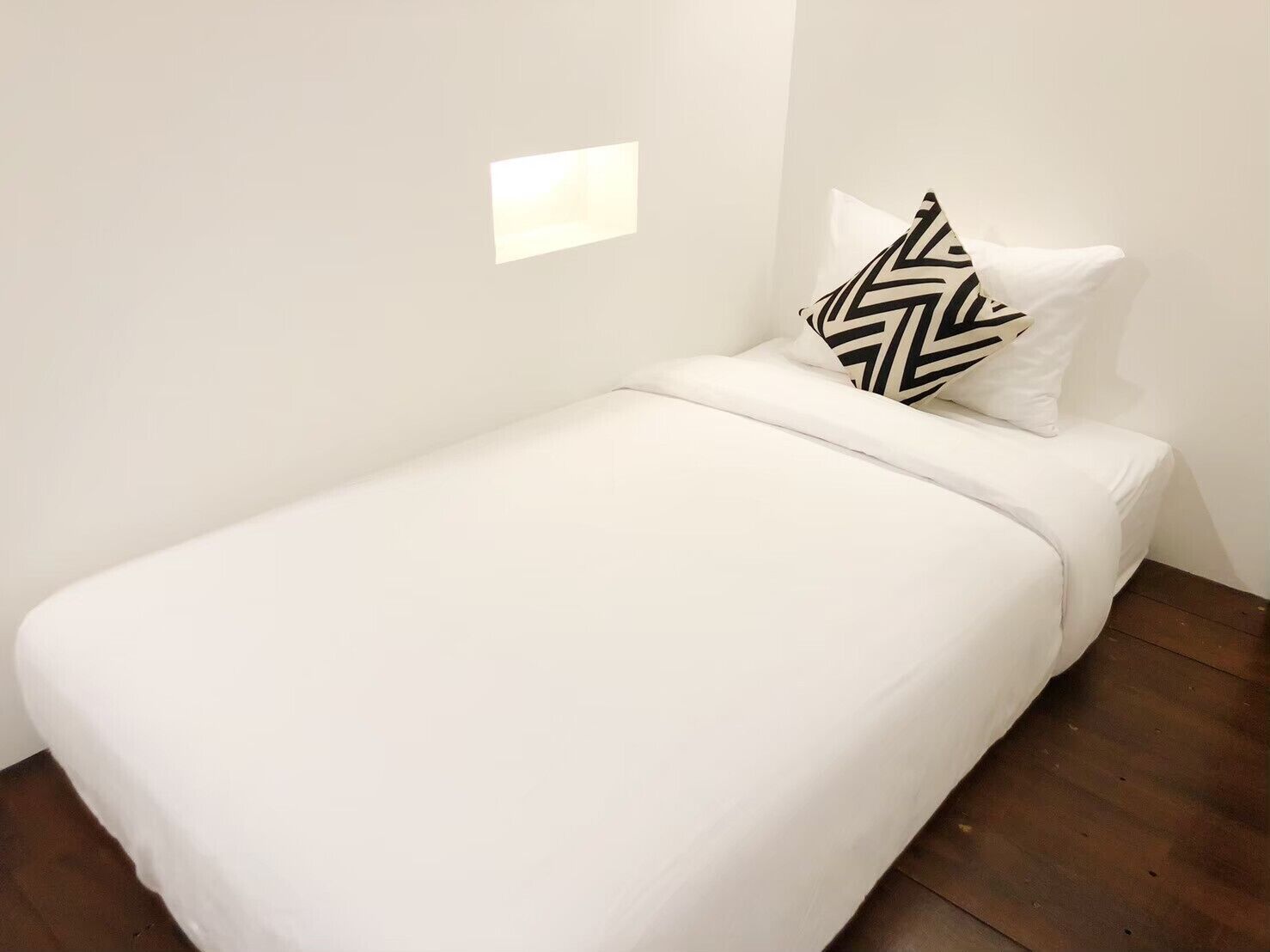 Ein kleines, ordentlich bezogenes Bett mit weißer Bettwäsche und einem schwarz-weißen Kopfkissen in einem minimalistischen Raum mit Holzboden und weißen Wänden.