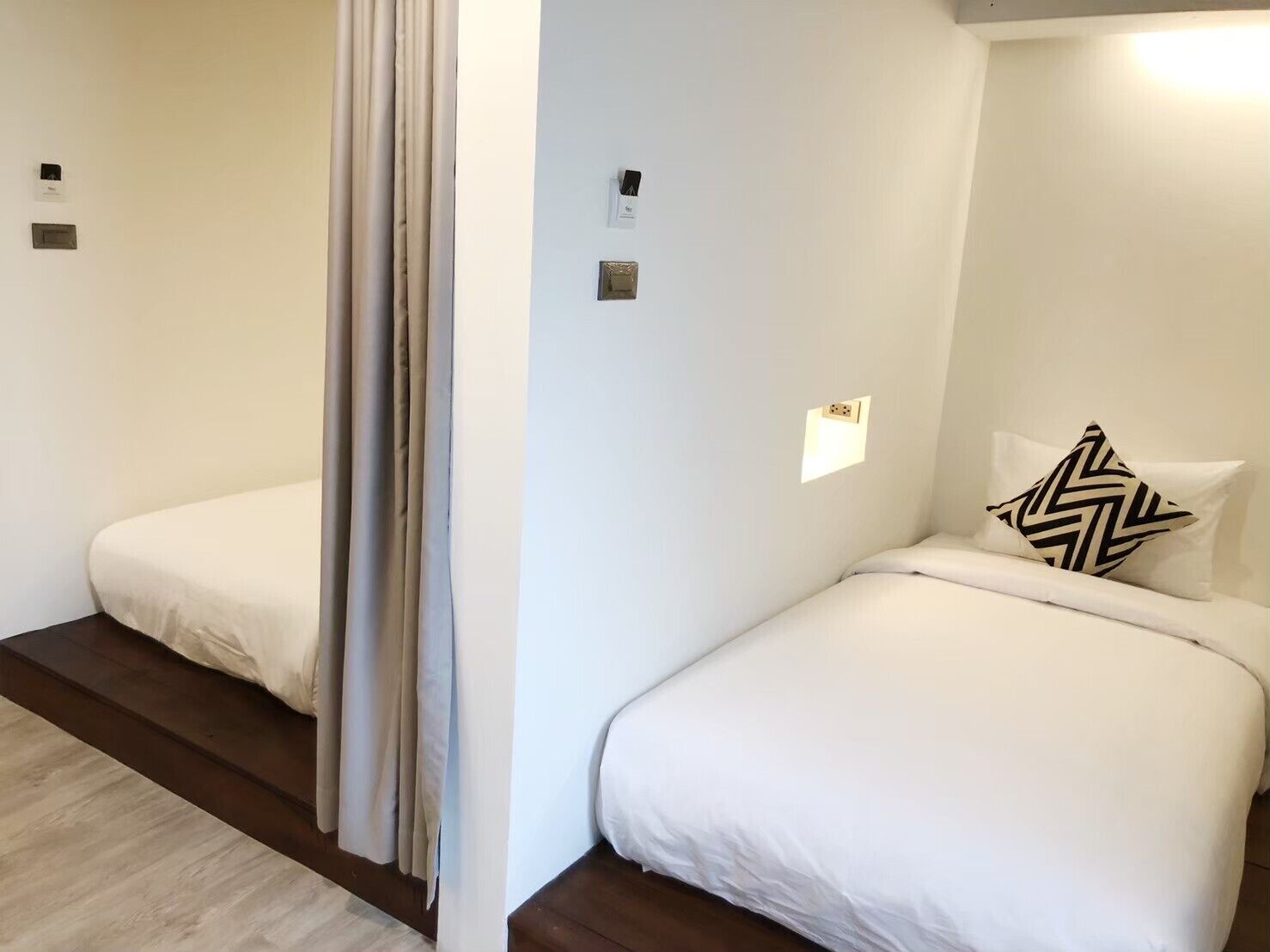 A hotel room with two beds, a wooden floor, and a white wall with a small window.