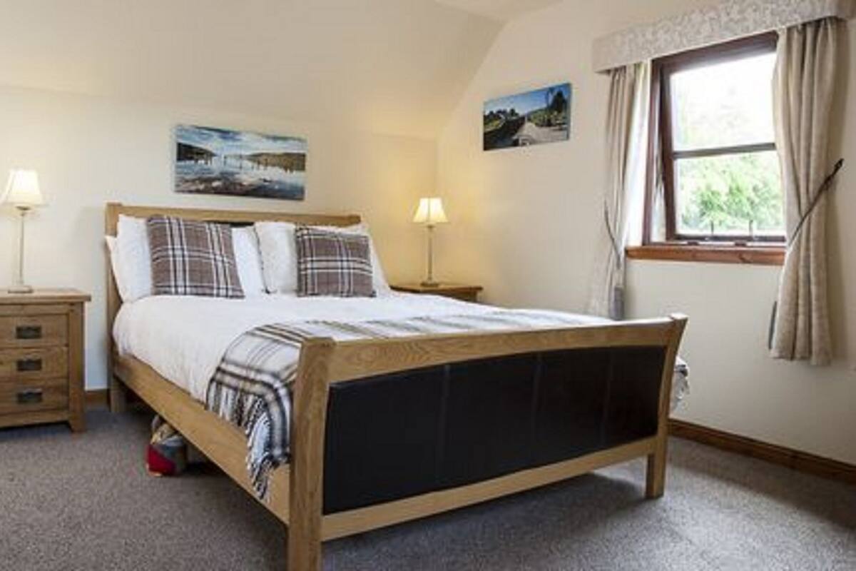 A bedroom with a wooden bed, two bedside tables with lamps, a window with curtains, and two wall-mounted pictures.