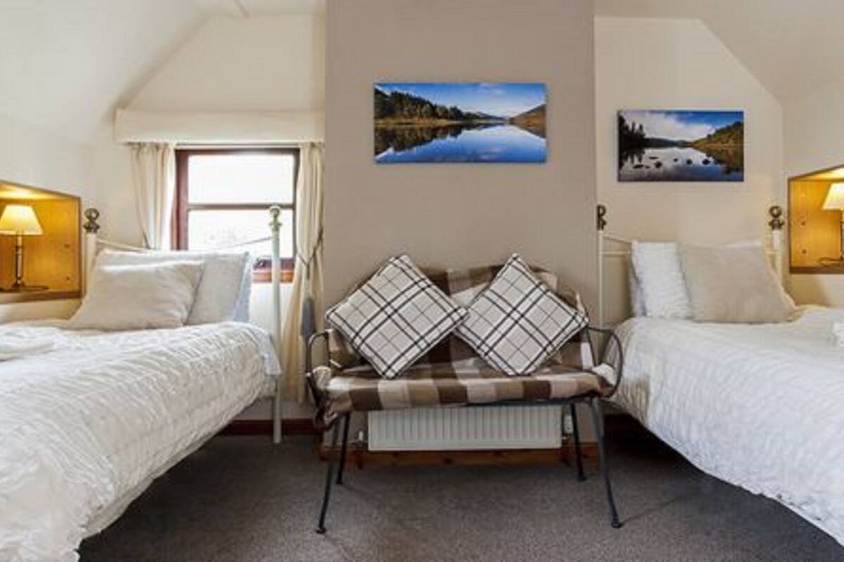 A hotel room with two single beds, a bench with plaid cushions, and two paintings of landscapes on the walls.