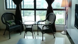 A room with two wicker chairs, a small table, a TV, and a view of a cityscape through large windows with dark curtains. - Lawang Suite 1 Bedroom Corner Apartment (Shah Alam)