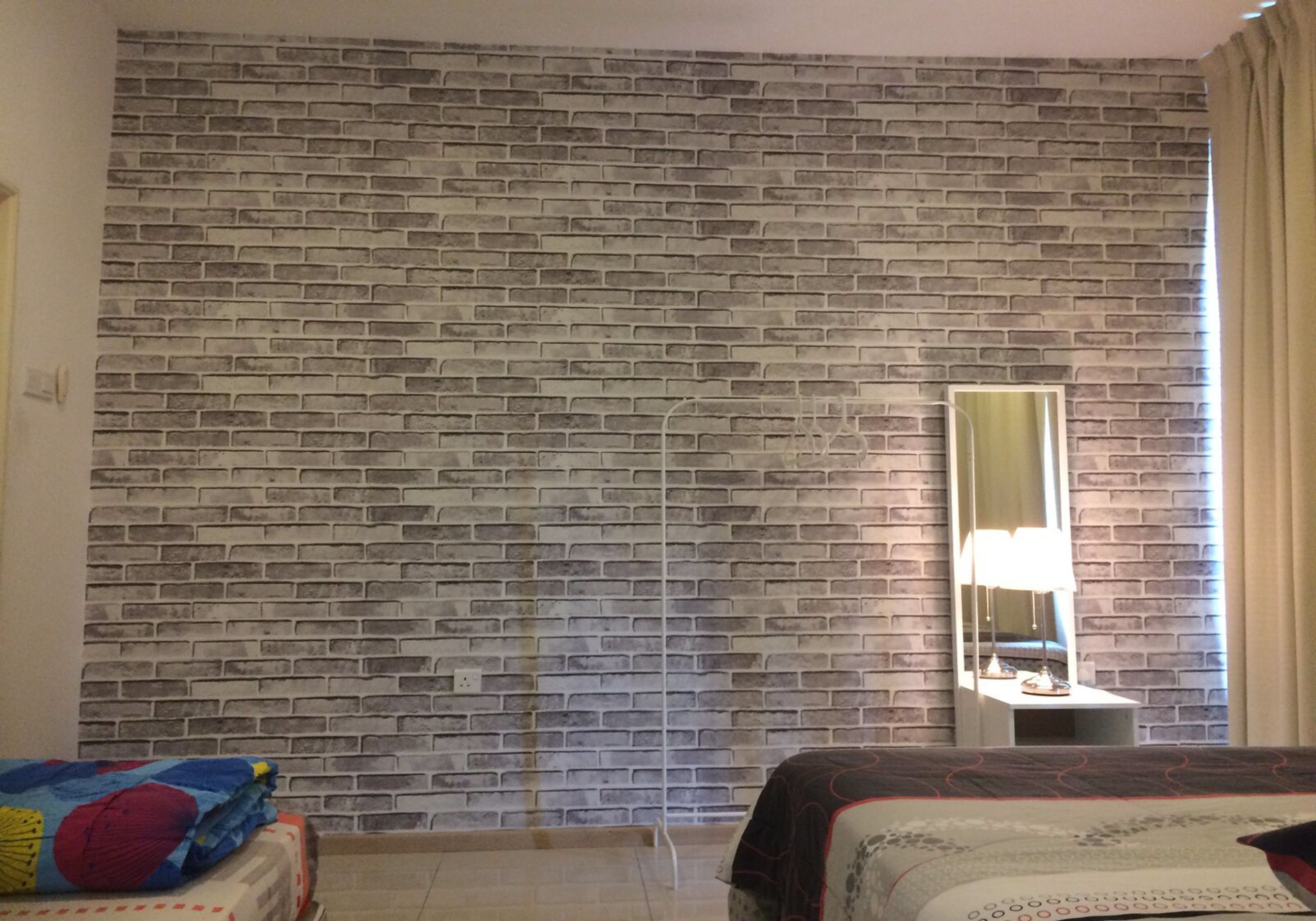 Une pièce avec un mur dont le motif imite des briques, un lit recouvert d’une couvre-lit à motifs, une table de chevet avec une lampe et un miroir.