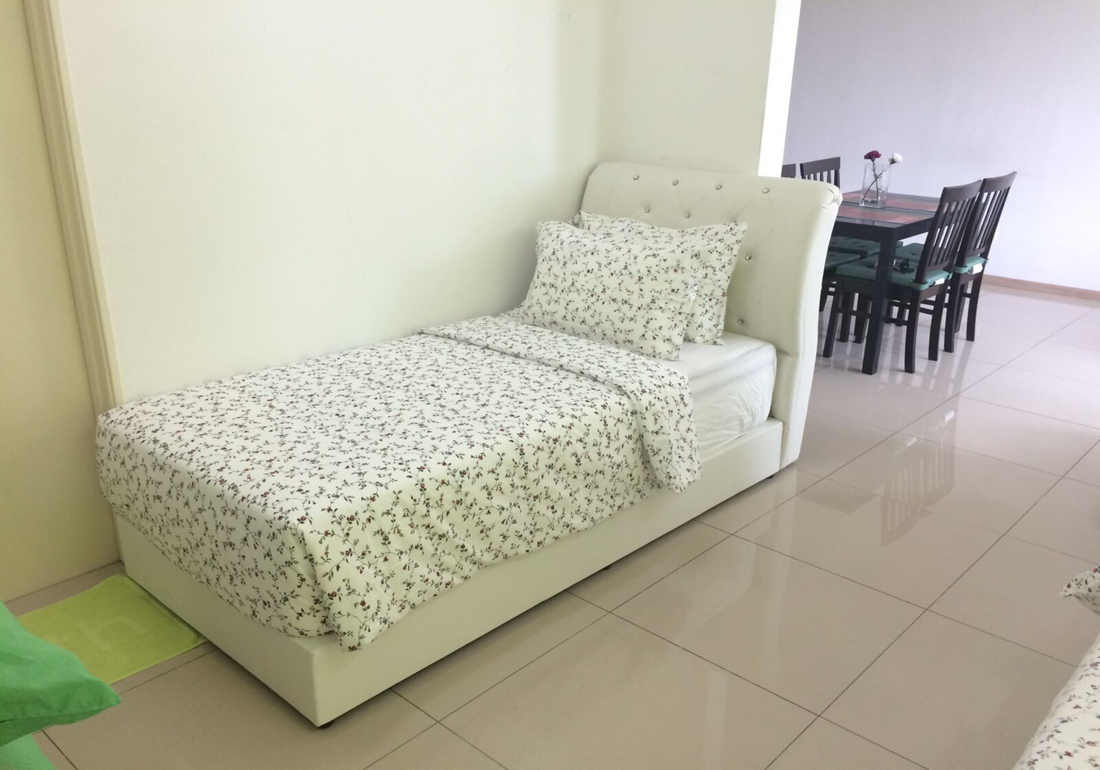 Un petit lit bien fait, avec une couvre-lit à motifs floraux et des coussins assortis, un tapis vert et un coin repas avec une table et des chaises à l’arrière-plan.