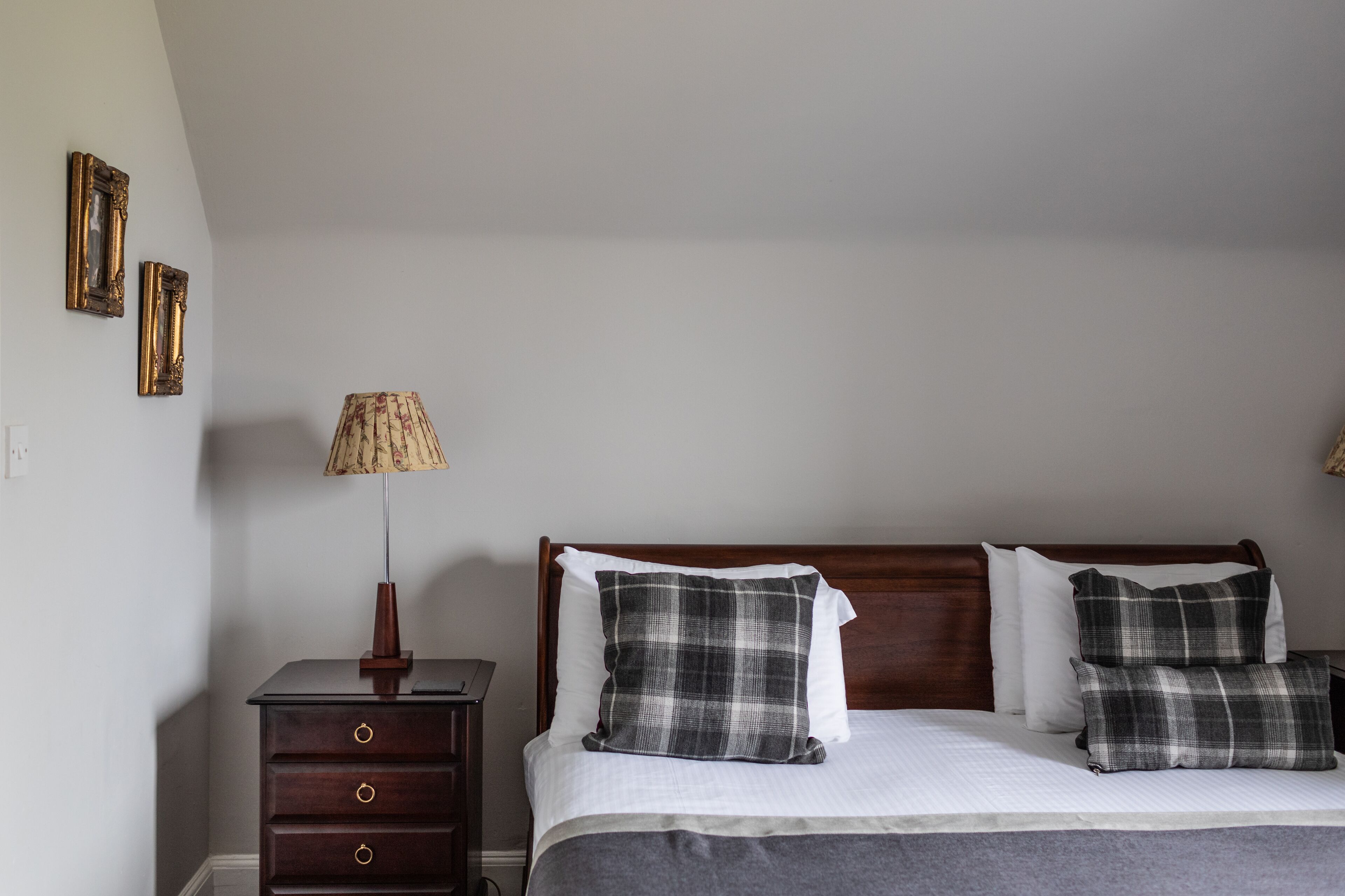 A neatly made bed with plaid pillows, a wooden nightstand with a lamp, and two framed pictures on the wall.