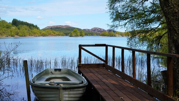 A wooden dock extending into a calm lake with a rowboat docked at the end. - Whitebridge Hotel (Inverness)