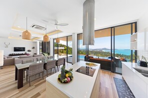 A modern kitchen with a central island, dining area with wicker chairs, and a large window overlooking a scenic view. - Samui Villa Bebe (Koh Samui)