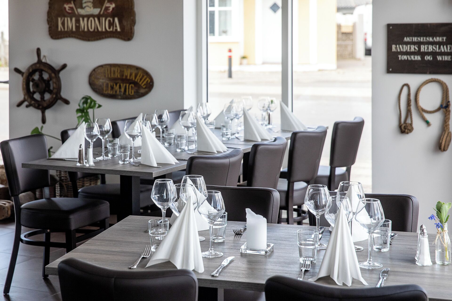 L’intérieur d’un restaurant, avec des tables dressées pour le service, une décoration murale sur le thème de la marine et une vue sur la rue.