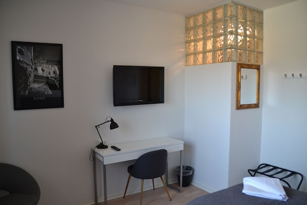 A bedroom with a bed, a desk, a chair, a TV, a mirror, and a window with a view of trees. - Thyborøn Hotel (Thyborøn)