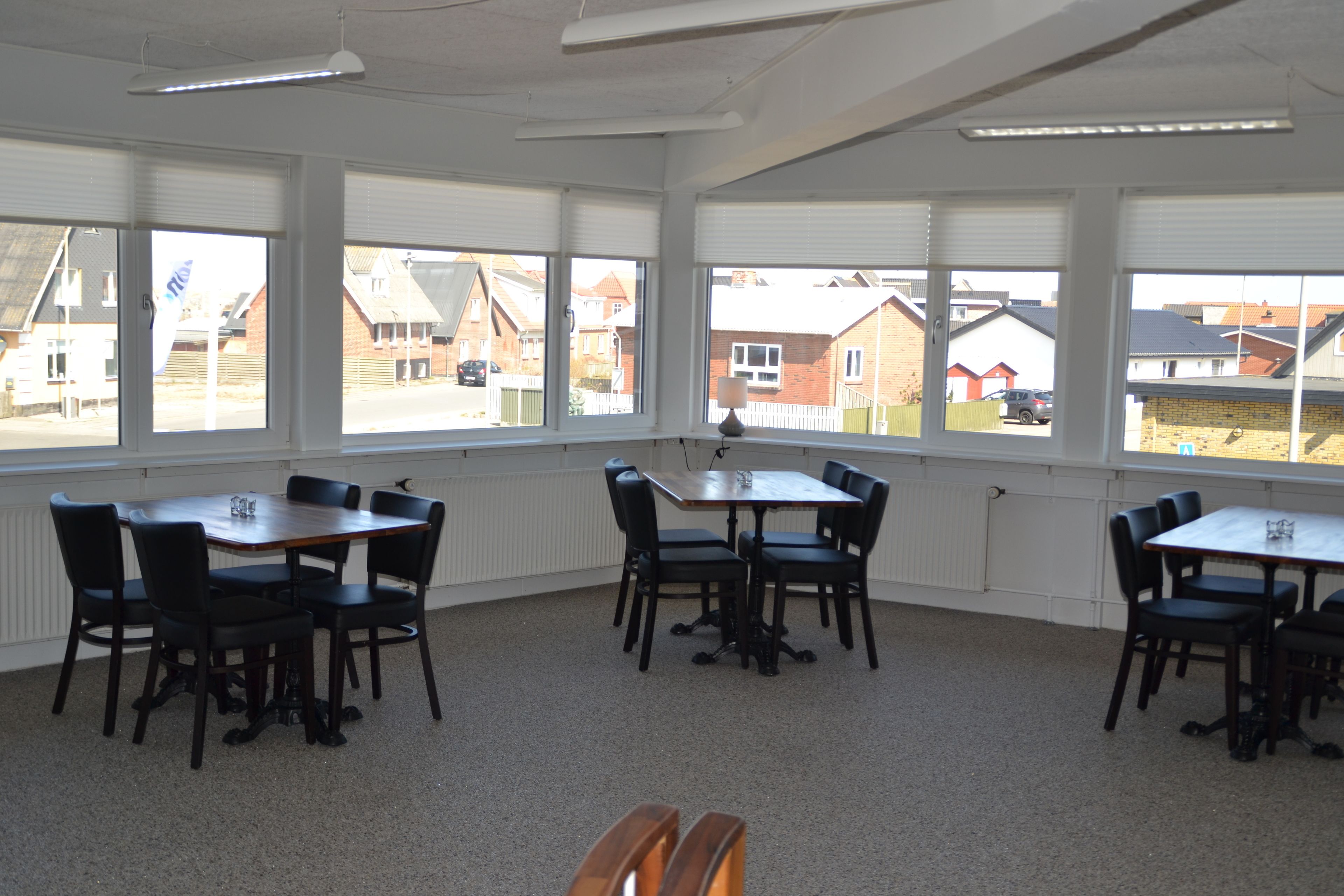 Un salón de conferencias con una gran mesa y sillas, ventanas amplias con persianas y vista a edificios residenciales.