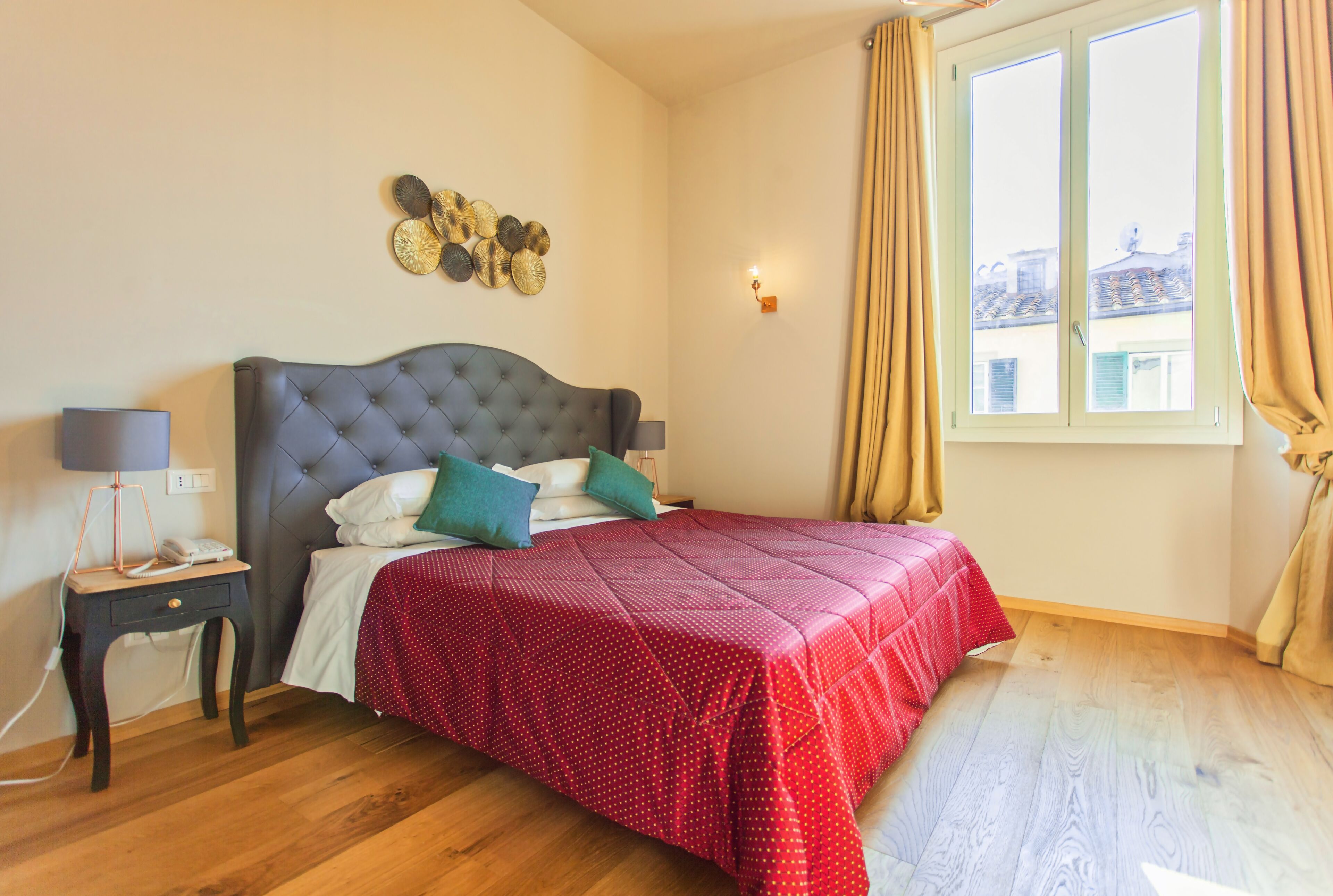 Une chambre à coucher avec une tête de lit capitonnée, une couvre-lit à pois rouges, une table de chevet avec une lampe et une fenêtre avec des rideaux.