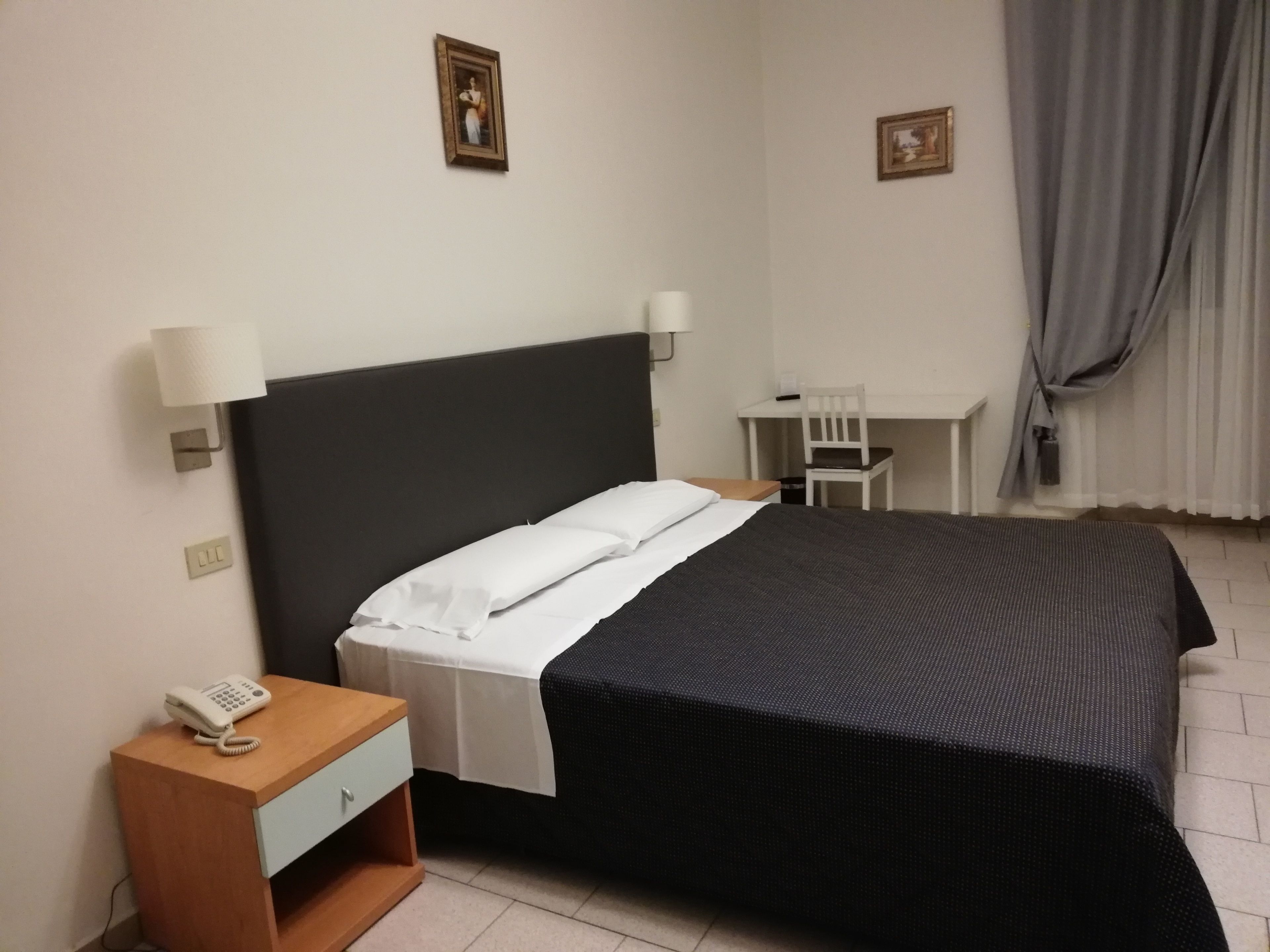 A hotel room with a bed, bedside table with a phone, a desk with a chair, and two wall-mounted lamps.