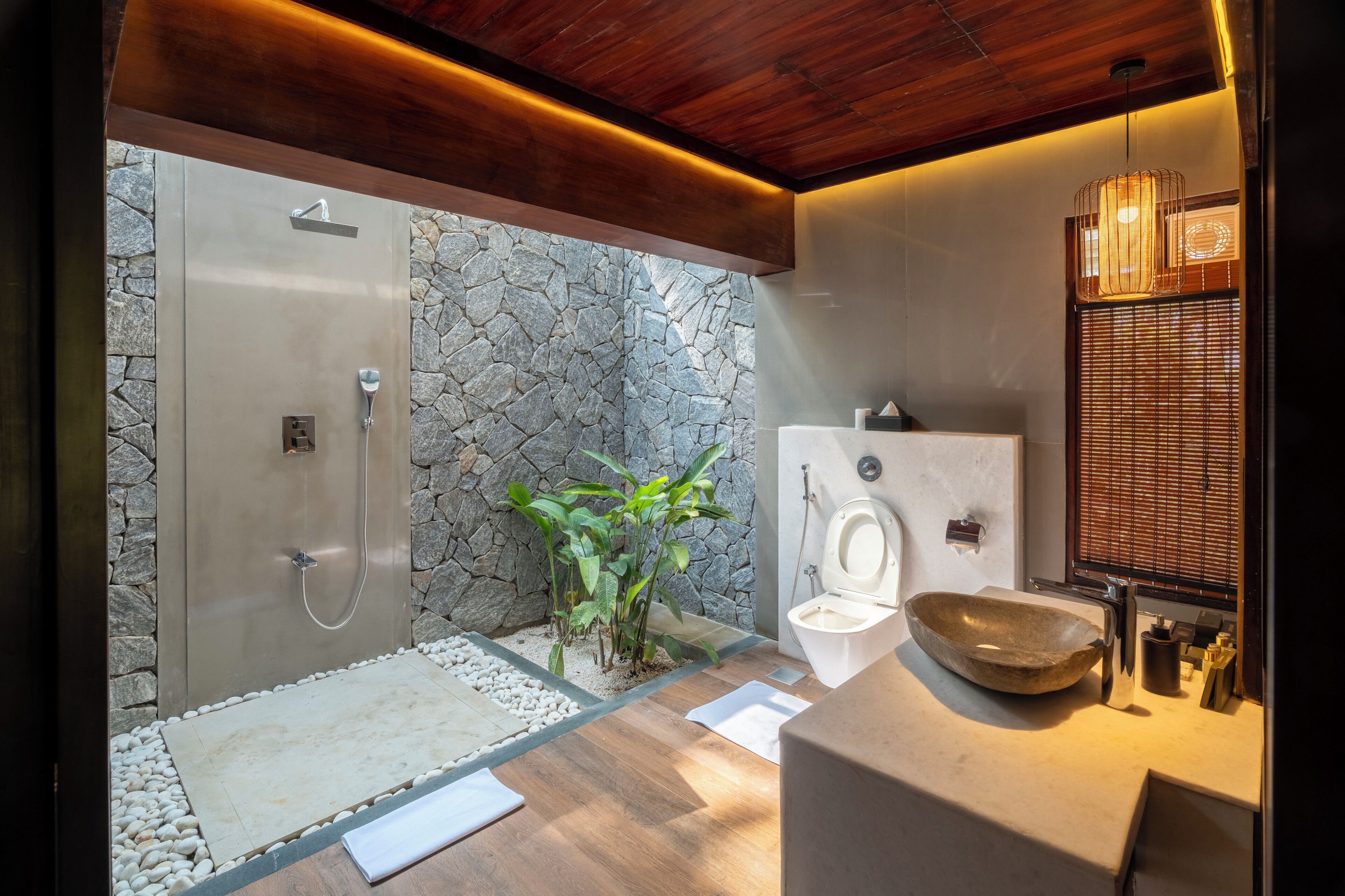 A modern bathroom with a stone wall, a wooden floor, a glass shower enclosure, a toilet, and a sink with a bowl.