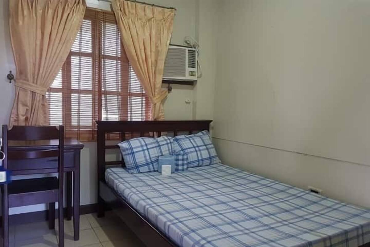 A small, single-bed room with a wooden headboard, a desk with a chair, beige curtains, and an air conditioning unit on the wall.