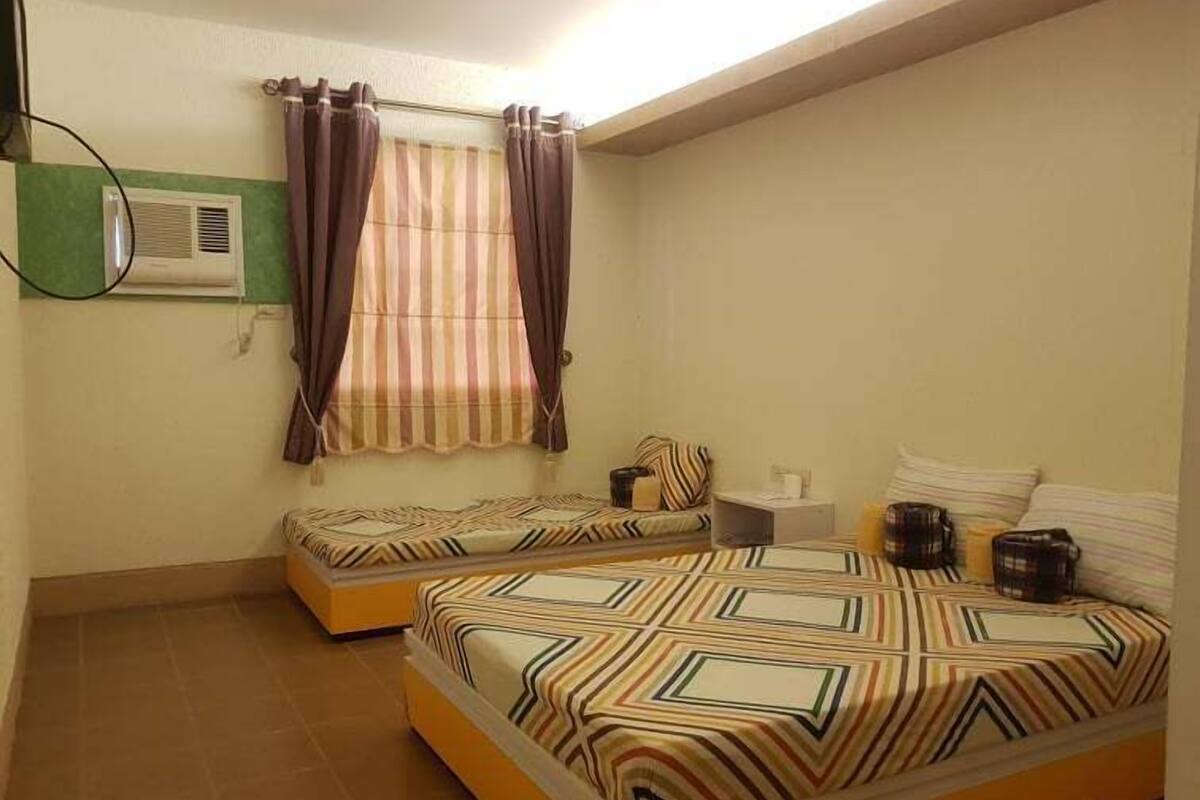 A small, well-lit bedroom with two beds, a window with curtains, a bedside table, and a wall-mounted air conditioner.