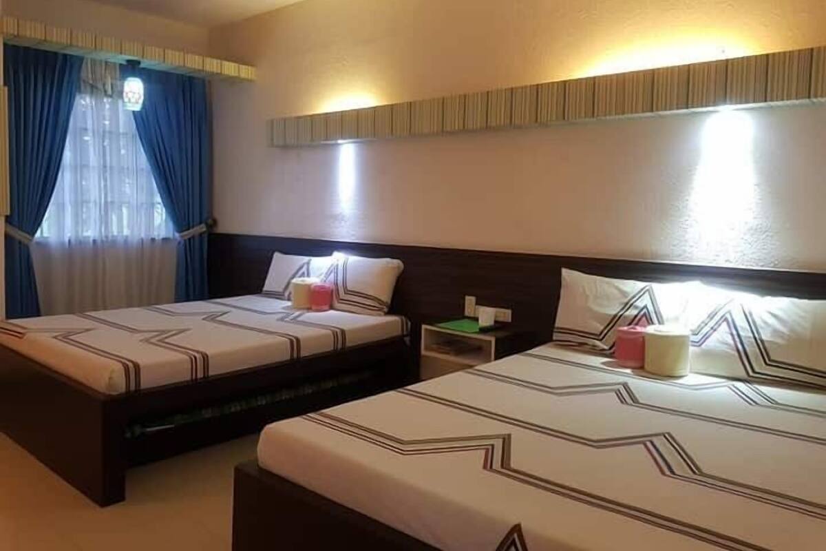 A hotel room with two beds, each with a headboard and pillows. There is a nightstand with a lamp and a small table with a green book and a white object.