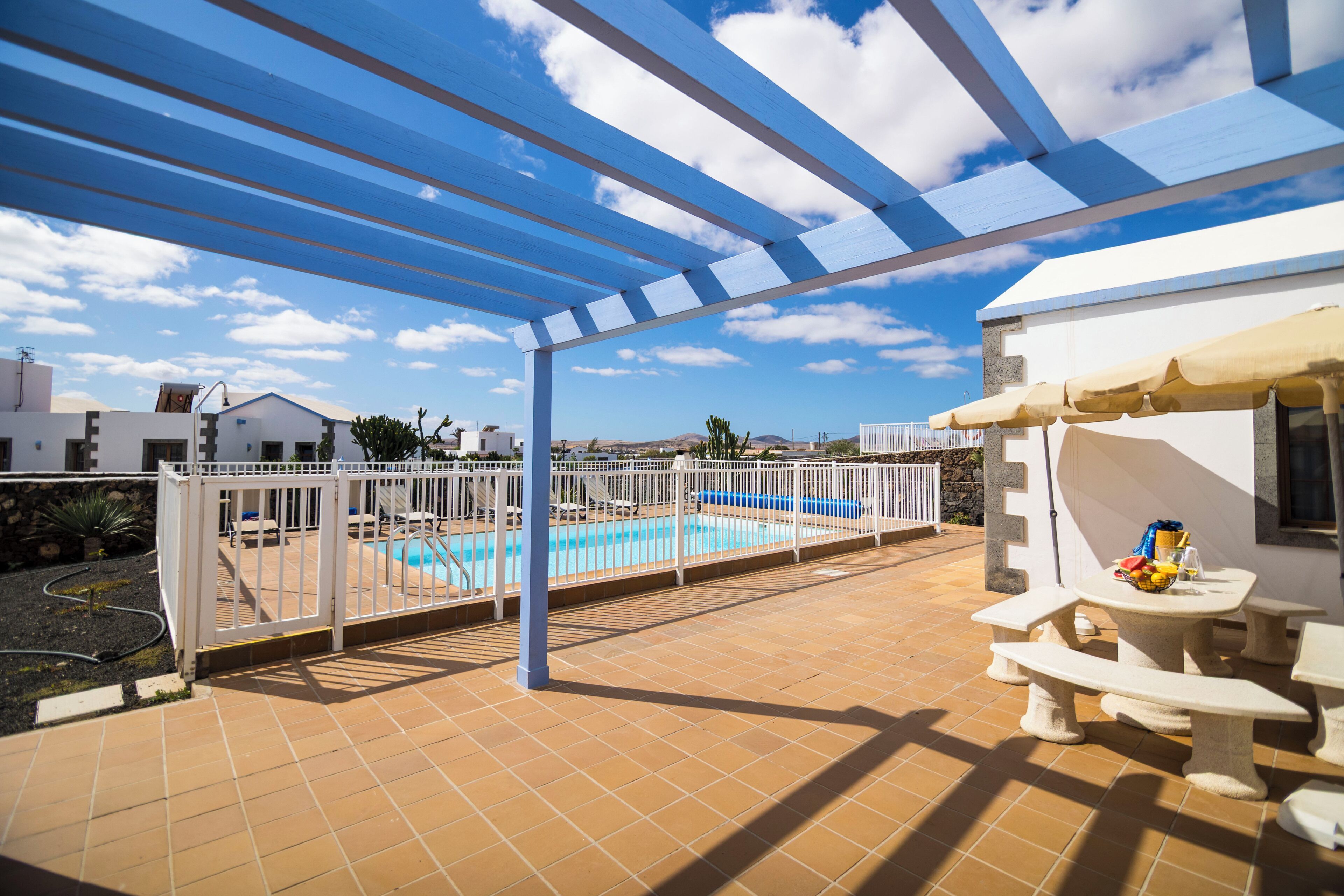 Un espacio exterior cubierto con piscina, mobiliario blanco y una pérgola.