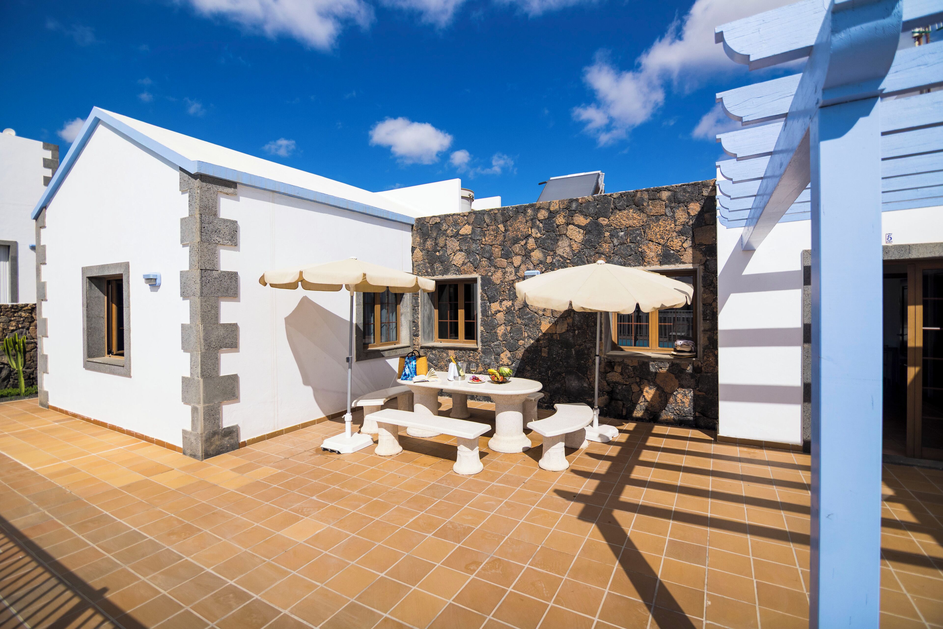 Un patio con una mesa y bancos, un muro de piedra y un edificio blanco con techo azul.