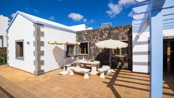 Un patio con una mesa y bancos, un muro de piedra y un edificio blanco con techo azul.