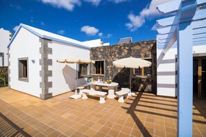 Un patio con una mesa y bancos, un muro de piedra y un edificio blanco con techo azul.