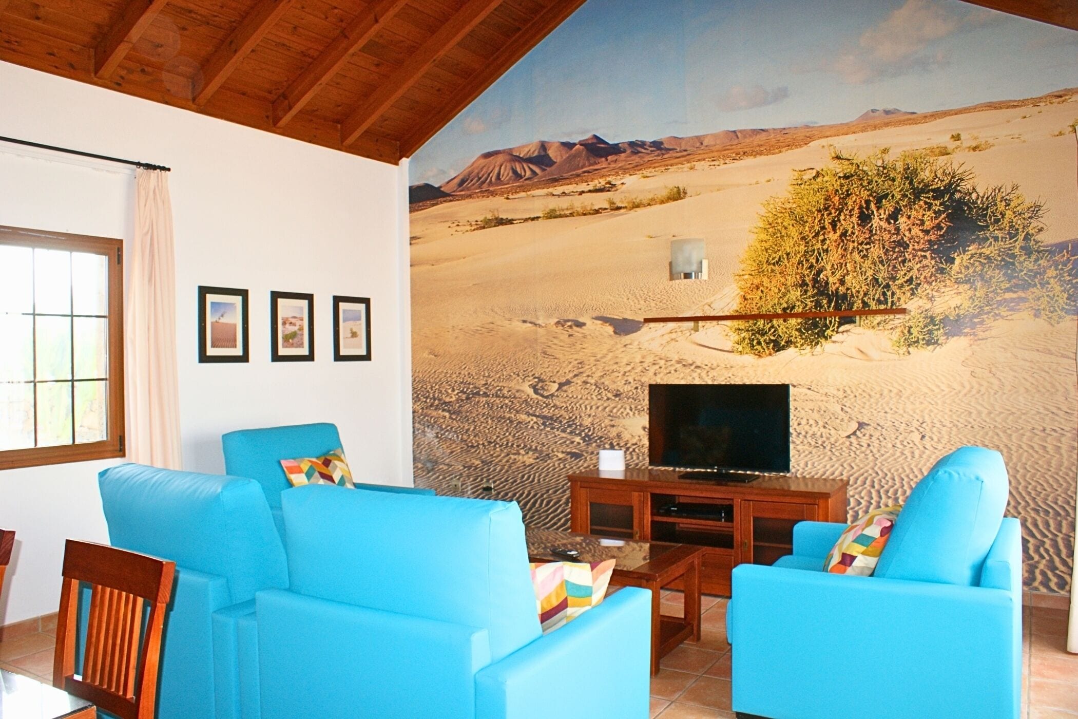 Una sala de estar con sofás azules, un televisor y un mural con motivos desérticos en la pared.