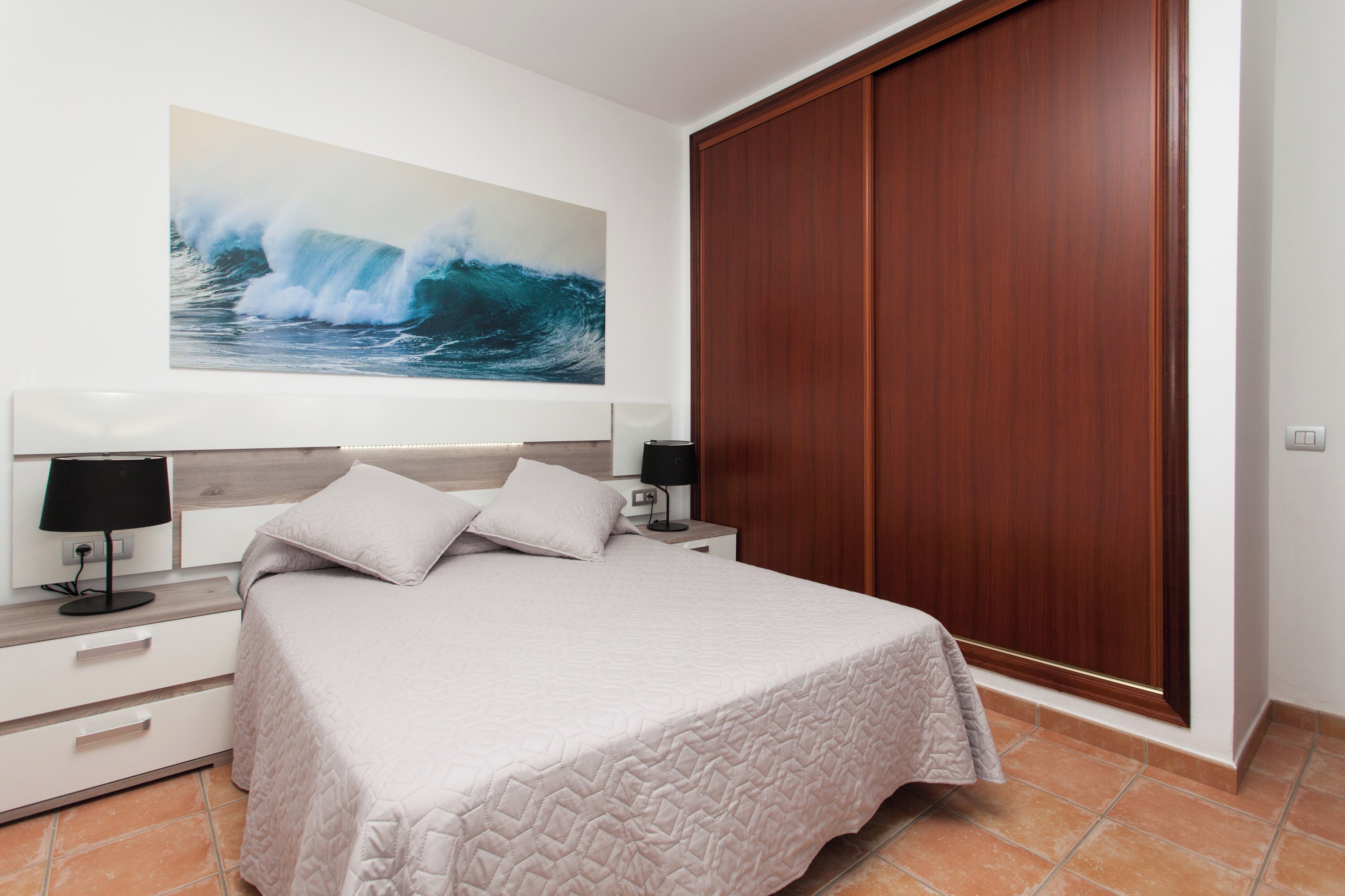 Un dormitorio con cama, mesitas de noche, una lámpara de pared, un armario de madera y un cuadro grande de una ola marina.