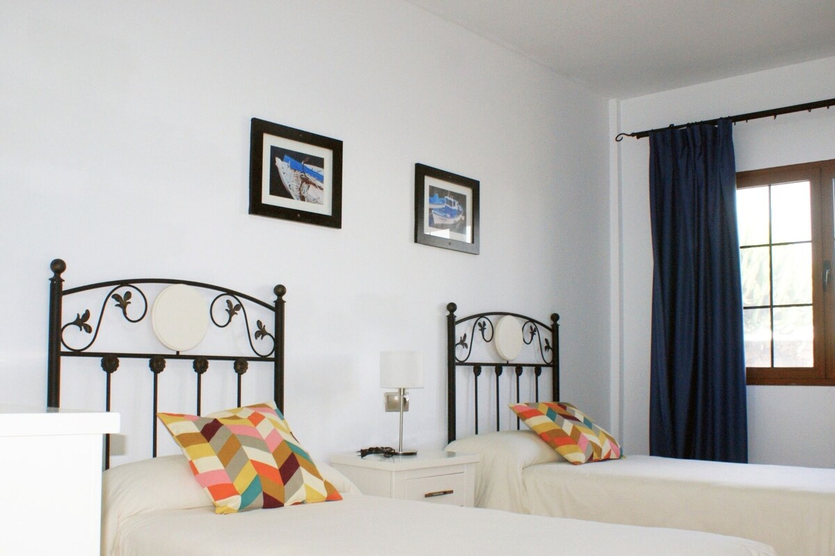 Una cama doble con cabecera de metal, una mesita de noche con lámpara y dos cuadros enmarcados en la pared.