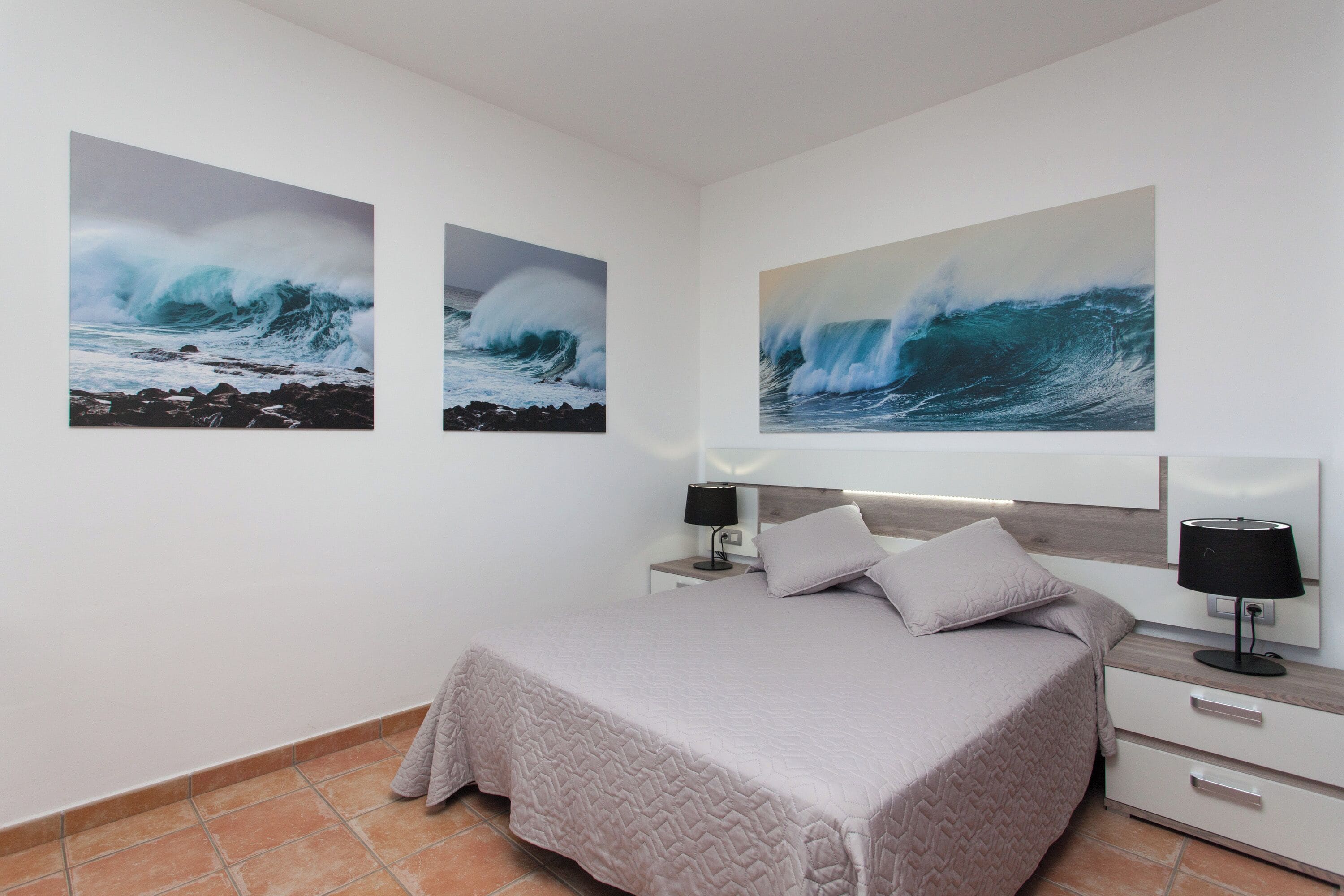 Un dormitorio con una cama, mesitas de noche, lámparas y tres grandes cuadros de olas en la pared.