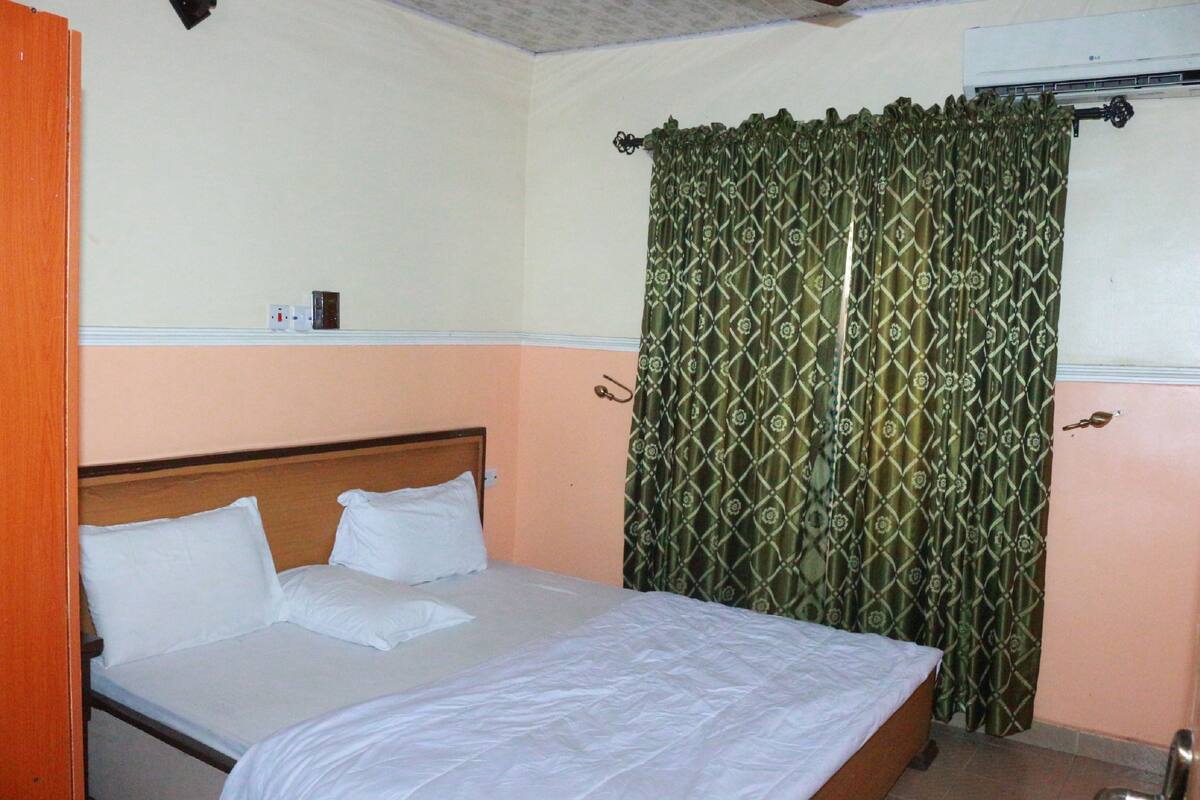 A single bed with white linens in a room with peach walls and a green patterned curtain.