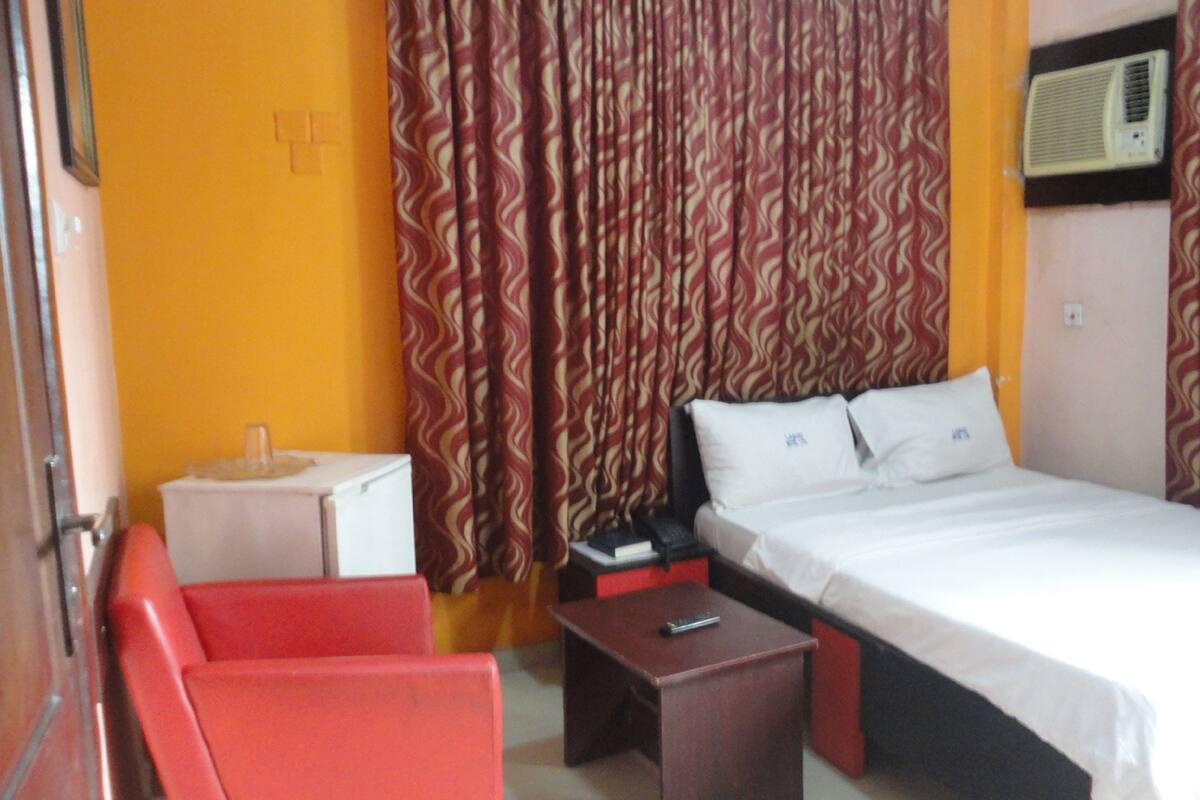 A hotel room with a bed, a red chair, a small table, and a bedside table.