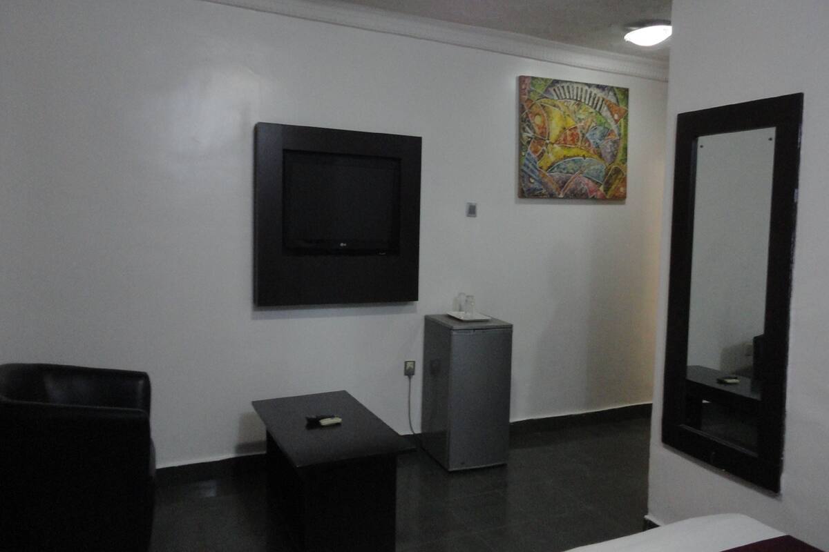 A room with a flat-screen TV mounted on the wall, a small table with a remote control, a grey cabinet, and a large mirror.