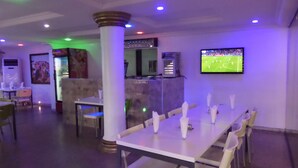 A restaurant with a long dining table, white chairs, and a TV screen displaying a sports event. - Labod Hotel (Ibadan)