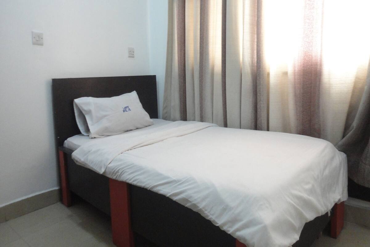 A single bed with a white mattress and pillow, a dark headboard, and a curtain partially drawn to the side.