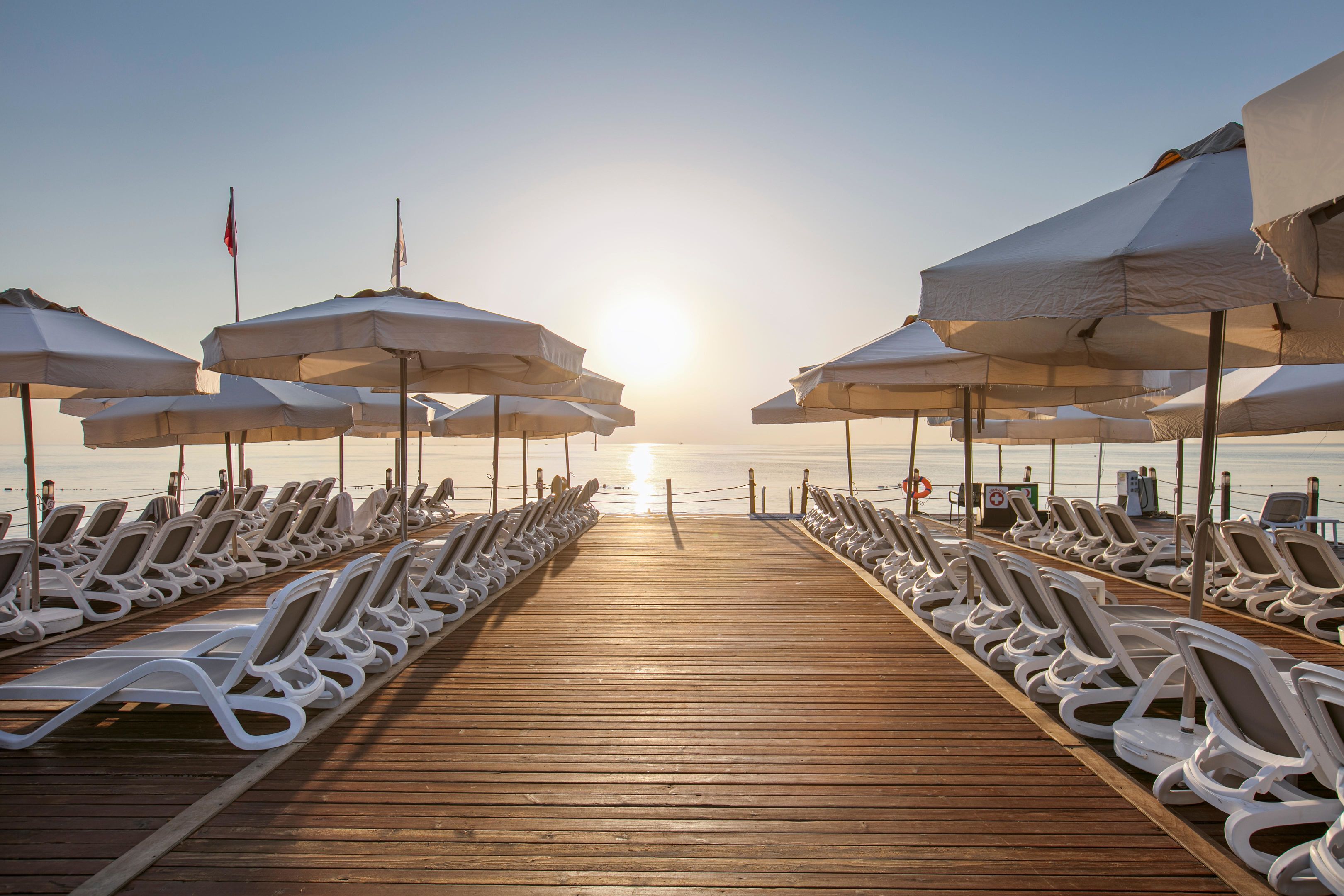 Eine Holzstege führt zu einem Strand mit Reihen aus weißen Liegestühlen und Sonnenschirmen.